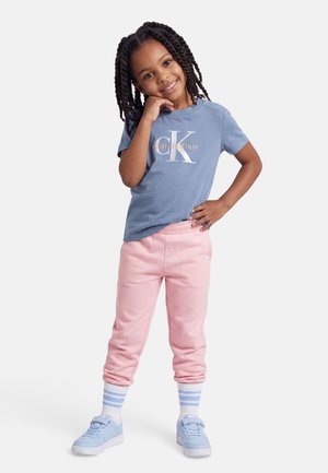 Χαμογελαστή κοπέλα με πλεγμένα μαλλιά στέκεται με το χέρι στη μέση, φορώντας μπλε μπλουζάκι Calvin Klein, ροζ παντελόνι, ριγέ κάλτσες και μπλε αθλητικά παπούτσια.