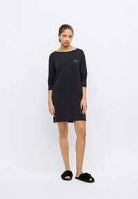 Black long-sleeve dress with a relaxed fit, featuring a round neckline and a small logo on the chest, paired with black fluffy slide slippers.