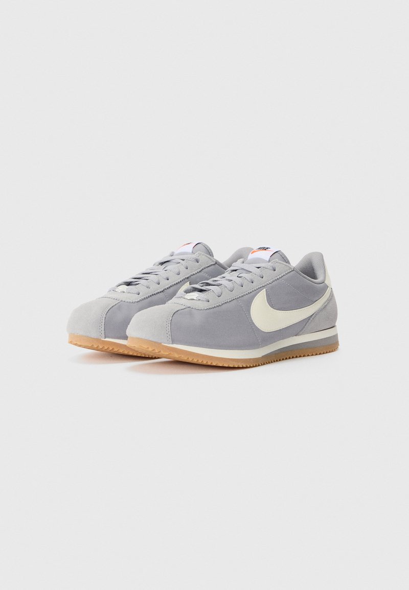 Graue Wildledersneaker mit einem weißen Nike-Swoosh, flachen Schnürsenkeln und einer Laufsohle aus Gummi. Verfügt über ein niedriges Design und eine runde Zehenform.