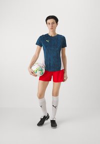 Sinise mustritega sportlik särk, punased lühikesed püksid, valged sokid logoga, mustad tossud; hoiab mitmevärvilist jalgpalli.