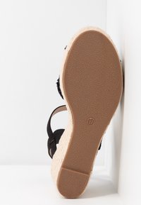 Semelle d’une sandale compensée avec une semelle en caoutchouc brun texturé, des brides noires et un talon compensé tissé beige clair, présentée sur fond blanc.