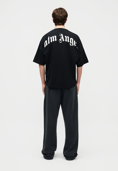 Uomo che indossa una t-shirt nera oversize con testo gotico bianco "Palm Angels" sul retro e pantaloni larghi di colore scuro, in piedi di schiena.