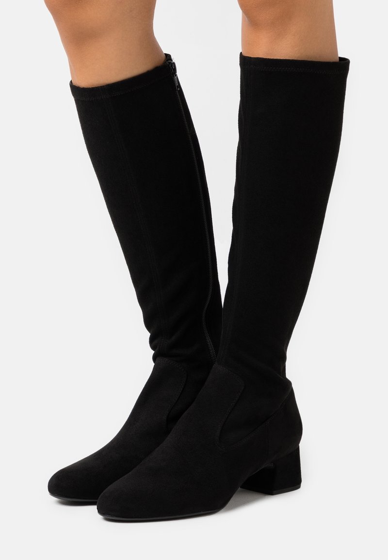 Bottes hautes en daim noir jusqu'aux genoux avec talons bloc et fermetures éclair latérales, portées sur des jambes nues, sur un fond clair uni.