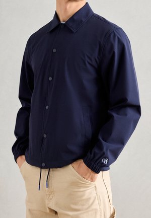 Mann trägt eine marineblaue Jacke mit Knöpfen und elastischen Bündchen sowie beige Hose, Hände in den Taschen, steht vor einem einfarbigen hellen Hintergrund.