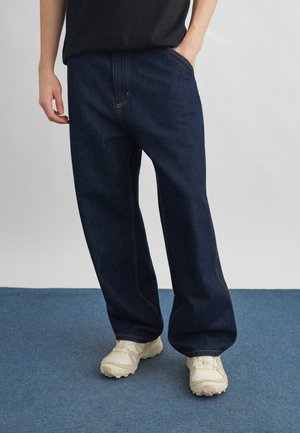 Άτομο που φορά χαλαρά σκούρα μπλε τζιν και λευκά αθλητικά παπούτσια, στέκεται σε μπλε χαλί με το ένα χέρι στην τσέπη.