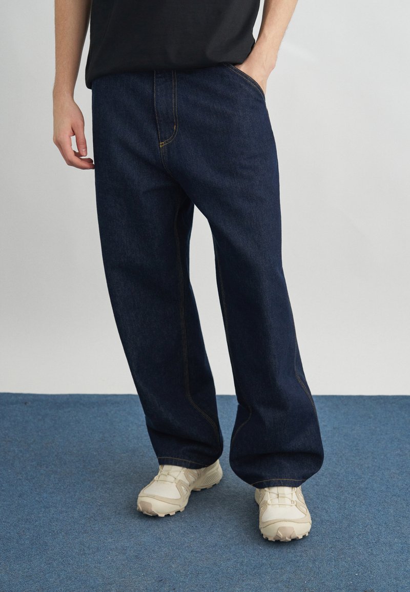 Osoba ubrana w luźne ciemnoniebieskie dżinsy i białe trampki, stojąca na niebieskim dywanie z jedną ręką w kieszeni.