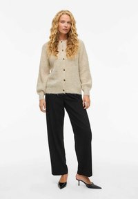 Μπεζ πλεκτή ζακέτα με μανίκια μπαλόνι και ξύλινα κουμπιά, σε συνδυασμό με μαύρο παντελόνι φαρδιάς γραμμής και μυτερά μαύρα τακούνια.