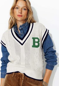 Gilet en tricot crème avec un patch vert en forme de "B", design en cable texturé, col en V et rayures navy. Porté par-dessus une chemise en denim bleu.