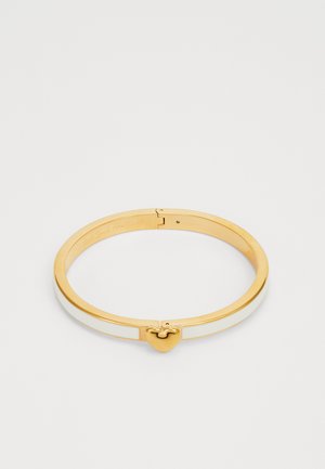 Nákar zlaté barvy s vkladem z bílé smaltu. Disponuje srdcovým zapínáním a hladkým, leštěným povrchem.