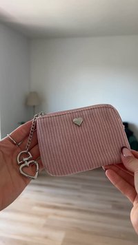 Borsa in tessuto rosa a coste con un accento a forma di cuore argentato e catena, dotata di chiusura con zipper. Forma compatta e rettangolare, superficie testurizzata.