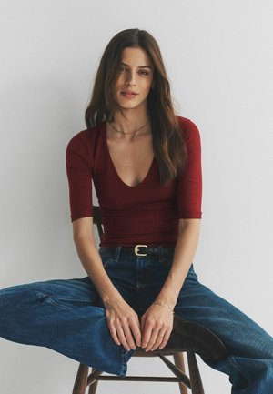 Bordový priliehavý top s hlbokým výstrihom do V, v páre so širokými modrými džínsami s rozšírenými nohavicami. Modelka sediaca na drevenej stoličke, ruky položené na kolenách.