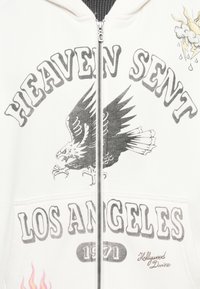 Felpa con zip bianca che presenta una grafica di un'aquila, il testo "HEAVEN SENT," "LOS ANGELES," e "1971" in nero. Include icone di fiamme e meteo.