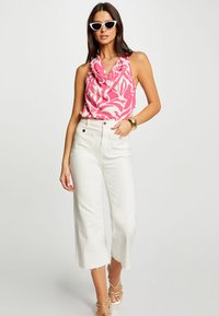 Top senza maniche rosa con motivo astratto bianco, abbinato a jeans bianchi a vita alta e a gamba larga. Gli accessori includono un bracciale d'oro e occhiali da sole bianchi.