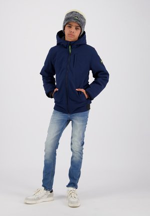Garçon portant une veste à capuche bleu marine, un bonnet en tricot gris, un jean bleu clair et des baskets blanches, se tenant les mains dans les poches devant un fond uni.