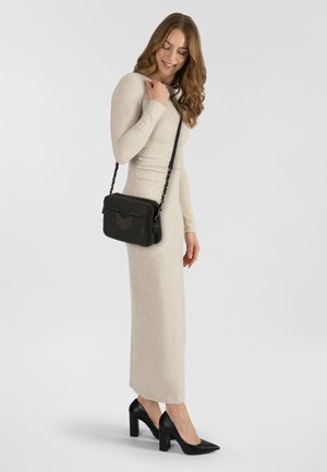 Schwarze Umhängetasche mit Nietenakzenten und Reißverschluss; verstellbarer Kettenriemen; kombiniert mit einem taillierten beige Strickkleid und schwarzen Absatzschuhen.