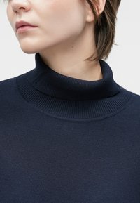 Tamnor blue pulover s turtleneck-ovim ovratnikom z ribasto teksturo, z usklajenim ovratnikom in gladkim telesom. Preprosta, minimalistična zasnova brez okrasnih elementov.