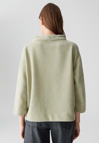 Frau von hinten gesehen, die einen hellgrünen, gerippten Pullover mit drei Viertelärmeln und dunkelgrauen Jeans trägt, vor einem schlichten Hintergrund.