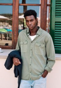 Jeune homme portant une chemise boutonnée vert clair et un jean clair, tenant une veste sombre, debout à l'extérieur près d'une fenêtre en bois et de volets verts.