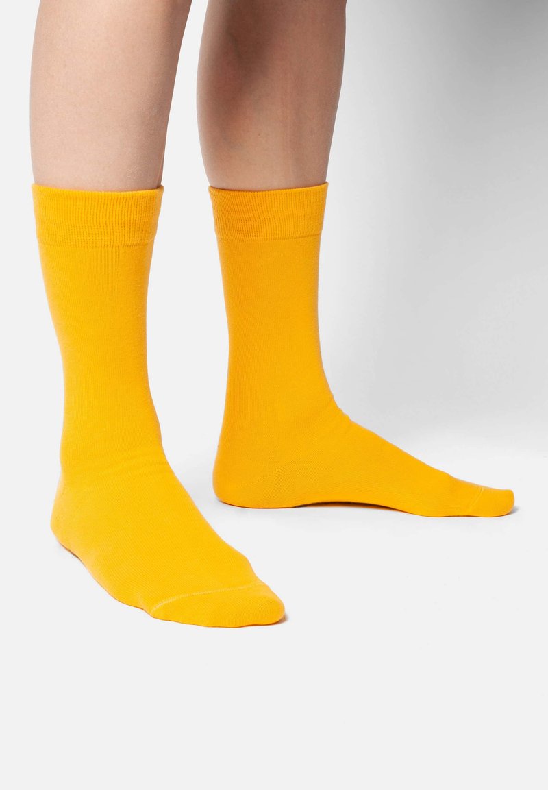 Gelbe Baumwollcrew-Socken mit geripptem Bündchen, die eine glatte Textur und eine knielange Passform aufweisen, auf den Füßen einer Person dargestellt.