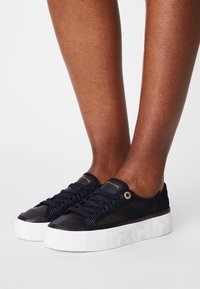 Tmavě modré tenisky s texturovaným svrškem, bílou platformovou podrážkou a lesklými detaily. Mají zlaté průchodky a zavazování na tkaničky.