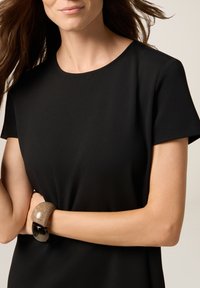 Schwarzes kurzärmeliges Kleid mit rundem Halsausschnitt und glatter Textur. Das Model trägt ein großes, zweifarbiges Armband an einem Handgelenk.
