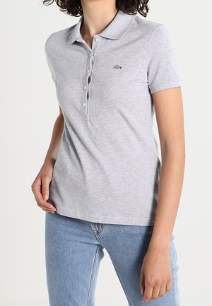 Vrouw draagt een lichtgrijs poloshirt met korte mouwen, knopen en een klein geborduurd logo, gecombineerd met blauwe denim jeans.