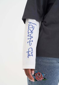 Zwarte T-shirt met korte mouwen, gelaagd over een witte blouse met lange mouwen, met blauwe gedrukte tekst. Donkerblauwe jeans met rode rozen borduursel.