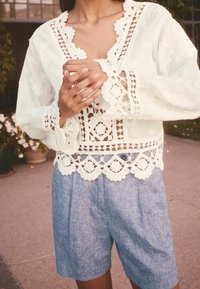 Bílá krajková blůza s jemnými výřezy a zvlněnými okraji, doplněná světle modrými lněnými šortkami s volným střihem a sklady.