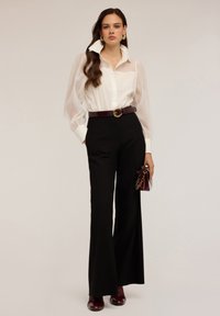 Blouse blanche en voile avec un col haut et des manches bouffantes, assortie à un pantalon noir taille haute, rehaussée par une ceinture et des accessoires bordeaux.