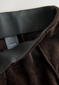 Brązowy dzianinowy wyrób z szerokim czarnym elastycznym pasem w talii. Metka głosi "NEXT". Tekstura jest prążkowana, a kolor jest głęboki brązowy.