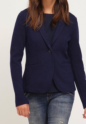 Kvinde iført en tætsiddende mørkeblå blazer med en enkelt knap, kombineret med en navy top og slidte blå jeans.