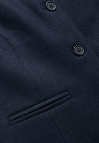 Námornícky modrý vlnený kabát s hladkou textúrou, ktorý má dva čierne gombíky a jednu náprsníkovú kapsu na ľavej strane.