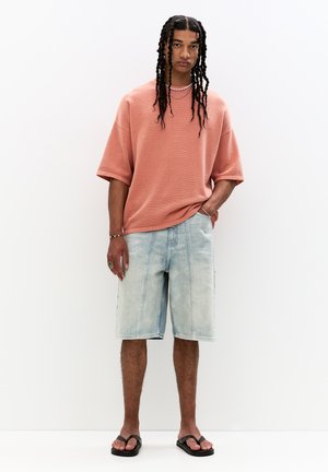 T-shirt in maglia di colore ruggine oversize con maniche corte, abbinata a pantaloni shorts di denim blu chiaro. Infradito neri; modella con collane a strati.