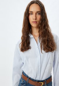 Camisa de botones a rayas azul claro con mangas largas y detalles de encaje en un lado, combinada con un cinturón marrón con tachuelas y jeans de mezclilla.