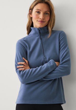 Vrouw met over elkaar geslagen armen, gekleed in een blauw fleece pullover met een kwart rits, staand tegen een lichte muur.