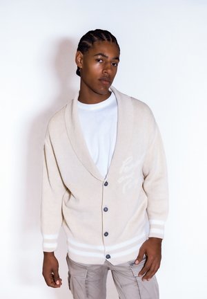 Beige Cardigan mit großem Schalkragen und Knopfverschluss. Besitzt cremefarbene horizontale Streifen an den Ärmeln und einen hellen Textdetail auf der Brust.