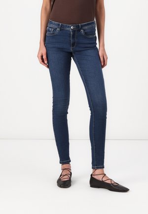 Skinny jeans in dark blue denim with a smooth texture, featuring a classic five-pocket design and contrasting stitching. Paired with black flats.