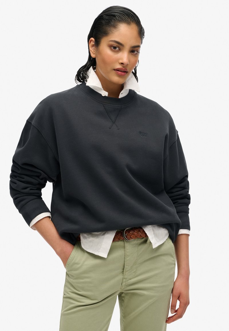 Felpa grigia scura con scollatura a girocollo, vestibilità rilassata e polsini a costine, indossata sopra una camicia bianca con colletto, abbinata a pantaloni verde chiaro.
