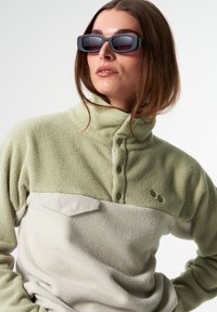 Frau mit einem zweifarbigen Fleece-Pullover mit hohem Kragen und Knöpfen sowie rechteckigen schwarzen Sonnenbrillen, die vor einem schlichten Hintergrund posiert.