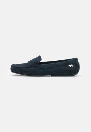 Mocassins en daim bleu marine avec un pli décoratif, des détails de couture et un accent de logo métallique sur le côté. Texture lisse et bout rond.