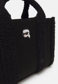 Fekete vászontáska plüss fekete fogantyúkkal, amelyen napszemüveget viselő arcot ábrázoló díszes címke található. Tartós szövetből készült.