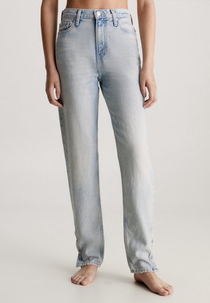 Personne portant un jean droit taille haute bleu clair avec des détails de boutons aux chevilles, debout pieds nus devant un fond uni.