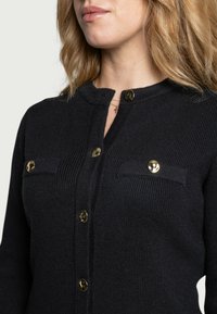 Navy Strickjacke mit goldenen Knöpfen, rundem Halsausschnitt und zwei Brusttaschen. Die Struktur ist gerippt, was ein strukturiertes Erscheinungsbild verleiht.