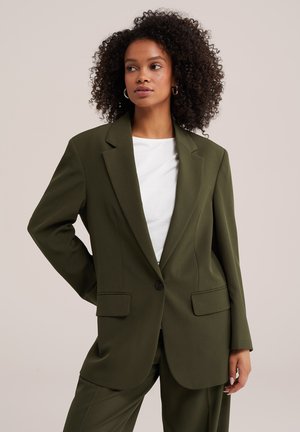 Donna con capelli ricci che indossa un blazer verde oliva oversize e pantaloni abbinati sopra una camicia bianca, in piedi con una mano sui fianchi.