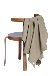 Krobelise mustriga oliivroheline tekk, mis on heledast puidust kõverate käetoega tooli peale heidetud, terava mustriga ja väikse sildiga äärel.