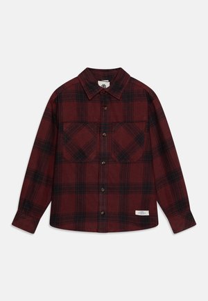 Chemise en flanelle à carreaux rouge et noir avec manches longues, devant boutonné, deux poches poitrine et une petite étiquette sur le côté inférieur. Texture douce.