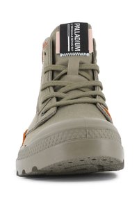 Zapatilla alta color verde oliva con suela de goma, parte superior de textil y acento naranja en contraste. Cuenta con una lengüeta etiquetada y ojales para los cordones.