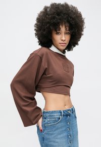 Sudadera marrón recortada con mangas largas y logo bordado; combinada con una falda vaquera de talle alto con botones en la parte delantera y lavado claro.