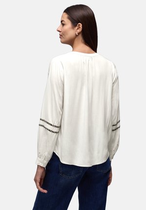 Vrouw met lang donker haar die een witte blouse met lange mouwen en zwarte rand aan de mouwen draagt, en donkerblauwe jeans, zijwaarts kijkend.