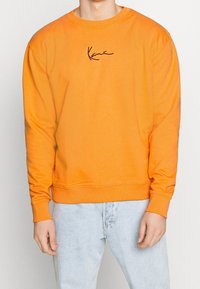 Orange bomullströja med rund halsringning och långa ärmar, med en svart signaturlogotyp på bröstet. Matchad med ljusa jeans.